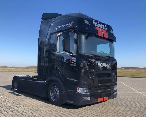 Nahaufnahme eines modernen, schwarzen Scania LKWs der BplusZ Group mit markanten orange-weißen Akzenten, geparkt auf einem gepflasterten Betriebshof vor klarem, blauem Himmel.